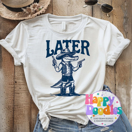White t-shirt with a graphic of a cowboy alligator holding guns and the word 'LATER' on a wooden surface.